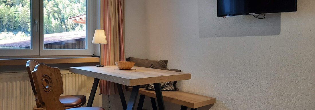A cozy dining area with a table, two chairs, and a television on the wall. Through the window, you can see green trees and nature outside. - Ferienhaus Anna - Sölden