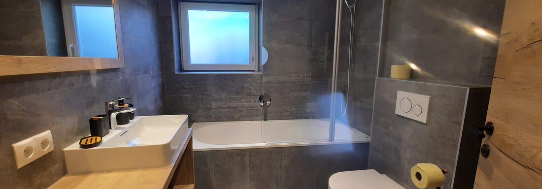 A modern bathroom with a bathtub, a sink, and a shower. The walls are in a gray stone look and there are wooden details. - Ferienhaus Anna - Sölden