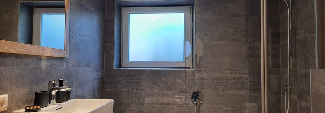A modern bathroom with gray tiles and a bathtub. The design is simple and bright, with a window for natural light. - Ferienhaus Anna - Sölden