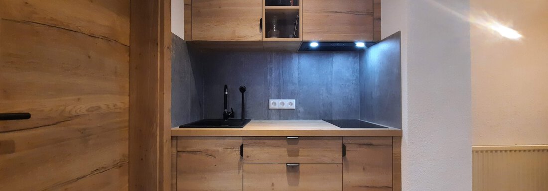 A modern kitchen corner with wooden cabinets and an elegant black workspace. The wall is subtly illuminated, creating an inviting atmosphere. - Ferienhaus Anna - Sölden