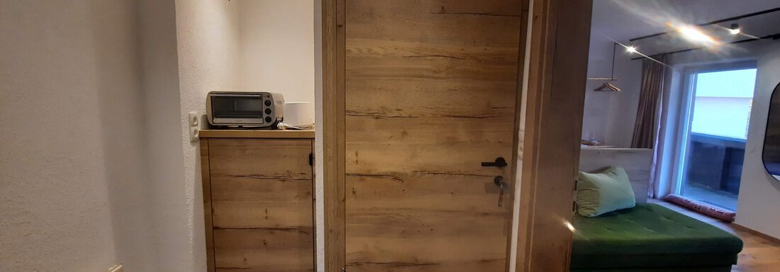A modern hallway with a wooden interior door and a small kitchen cabinet. On the right side, there is a cozy living area with a green sofa and plenty of natural light. - Ferienhaus Anna - Sölden