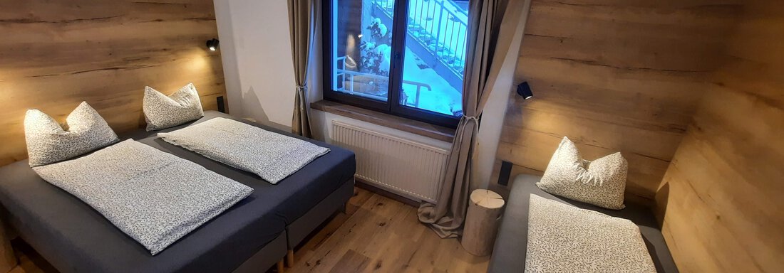 A modern bedroom with three beds and wooden paneling. Large windows provide plenty of natural light. - Ferienhaus Anna - Sölden