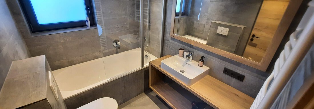 A modern bathroom with a bathtub, sink, and wooden shelf. It features a bright color scheme and large windows for natural light. - Ferienhaus Anna - Sölden
