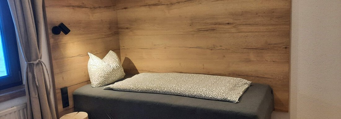 A modern bedroom with a simple bed and wooden paneling on the wall. There is a small table and a light source next to the bed. - Ferienhaus Anna - Sölden