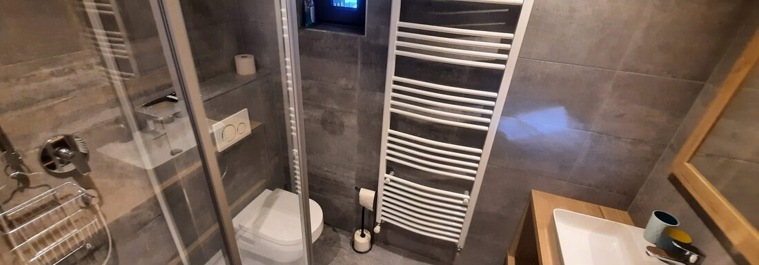 A modern bathroom with a glass-paneled shower and a sink. A towel heater is mounted on the wall and the floor has gray tiles. - Ferienhaus Anna - Sölden