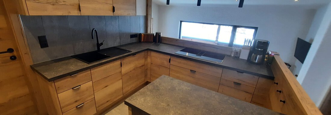 A modern wooden kitchen with an elegant counter and a large window. The kitchen elements are designed to be simple and functional. - Ferienhaus Anna - Sölden
