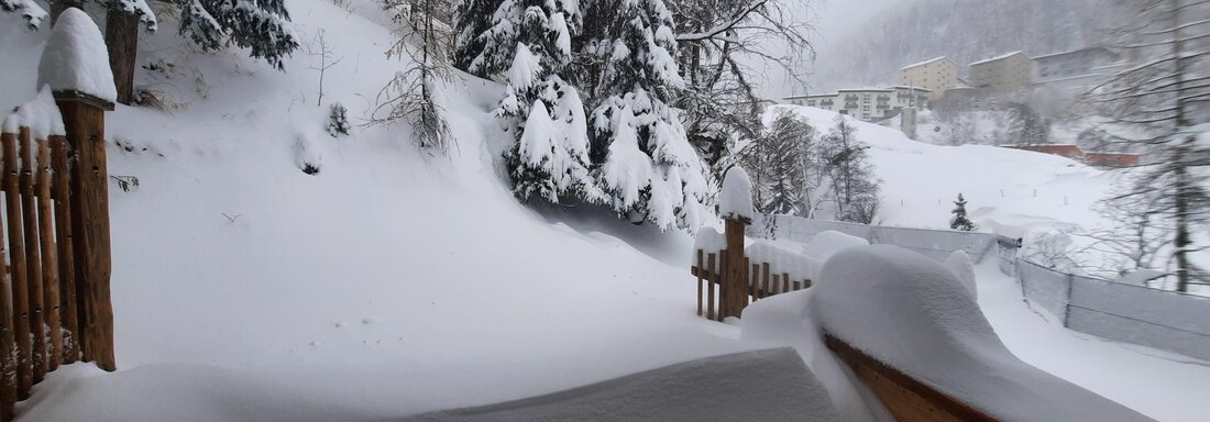 A snowy landscape with a lot of snow and tall trees. In the background, some buildings and a mountain can be seen. - Ferienhaus Anna - Sölden