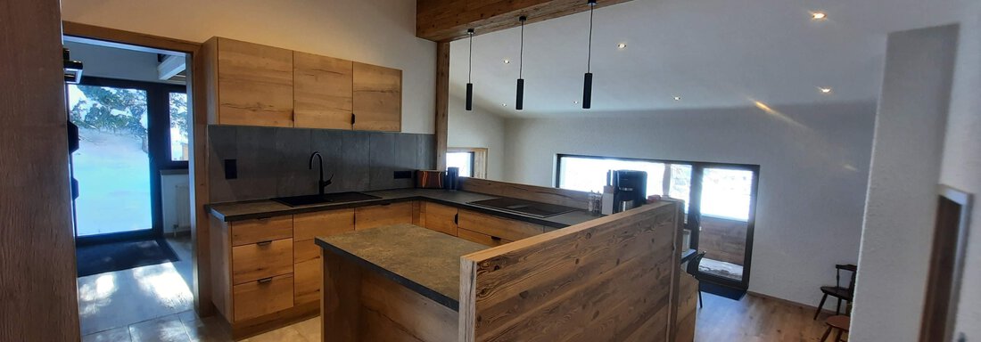 A modern kitchen with wooden cabinets and a large cooking island. Large windows provide plenty of daylight in the room. - Ferienhaus Anna - Sölden