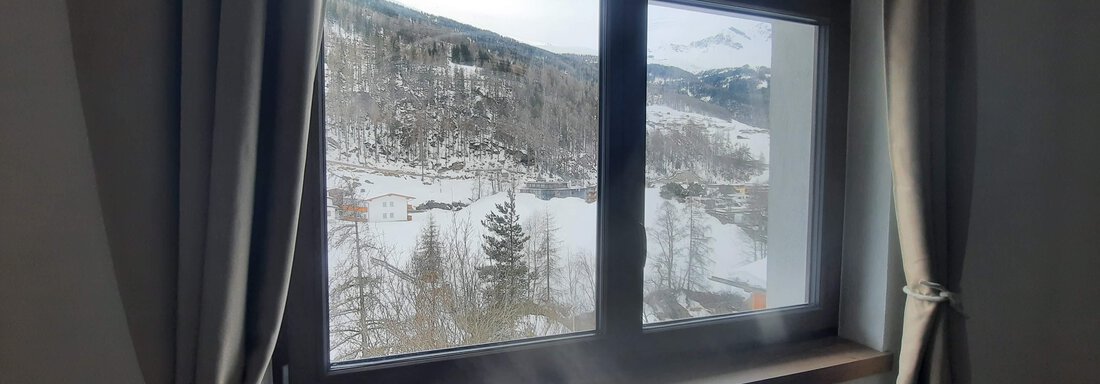 A window with a view of snow-covered mountains and trees. The curtains are light and the room feels cozy. - Ferienhaus Anna - Sölden