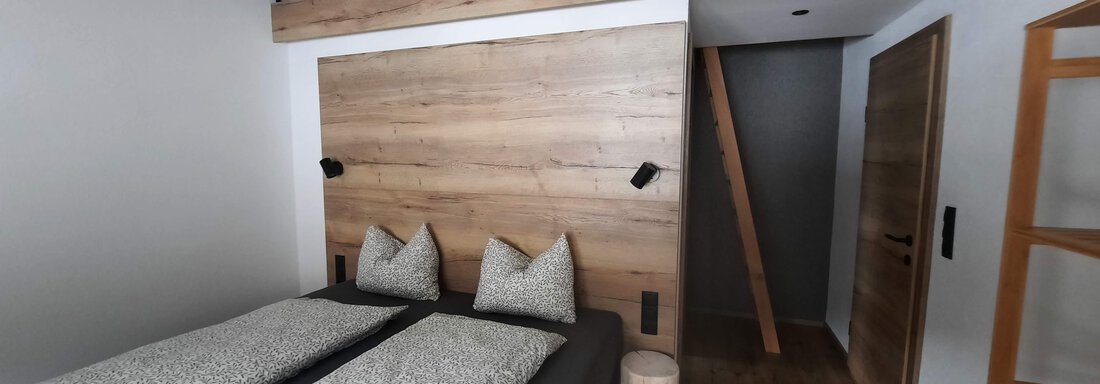 A modern bedroom with a double bed and wooden paneling. Above, there is a recessed area that is separated by a railing. - Ferienhaus Anna - Sölden