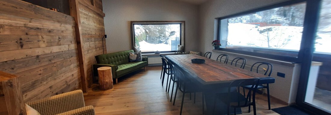 A modern and cozy dining room with a long table and a green sofa. Large windows offer a view of the snow-covered landscape. - Ferienhaus Anna - Sölden