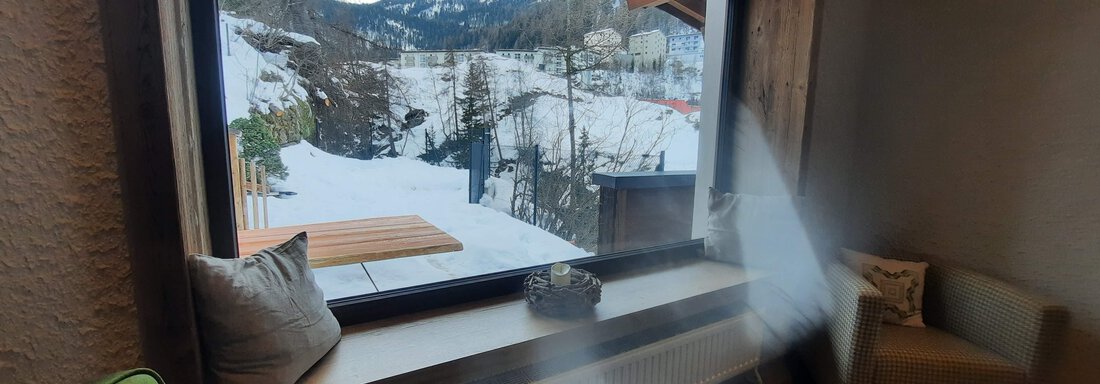 A cozy room with windows that offer a view of a snow-covered landscape. Sun rays stream through the window, illuminating the room. - Ferienhaus Anna - Sölden