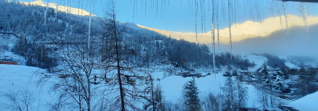 A snowy landscape with trees and mountains in the background. The sun rises behind the mountains and icicles hang from a roof. - Ferienhaus Anna - Sölden