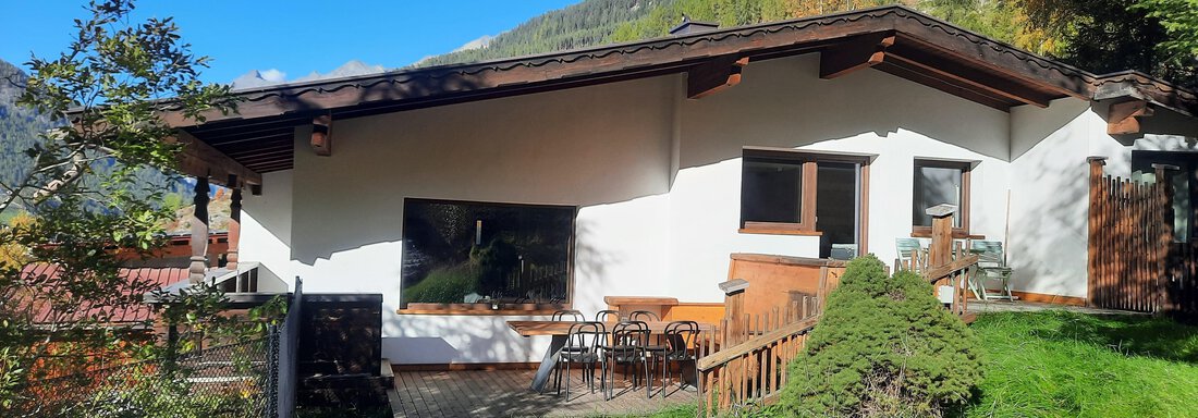A modern house surrounded by green grass and trees. In the background, mountains are visible under a clear blue sky. - Ferienhaus Anna - Sölden