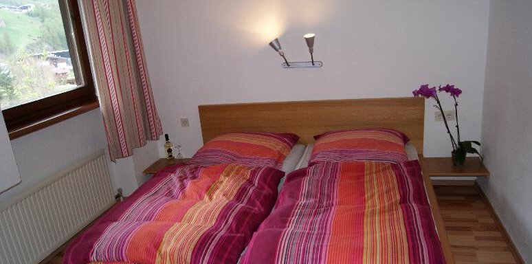 A cozy bedroom with a double bed and colorful bedspreads. Next to the bed is an orchid on the nightstand and a window lets in natural light. - Ferienhaus Anna - Sölden