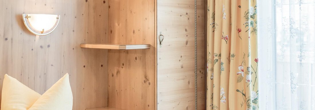 A cozy bedroom with a wooden wall and a large window. The yellow curtains and the decorative cushion create an inviting atmosphere. - Ferienhaus Apart Sonnzeit - Sölden