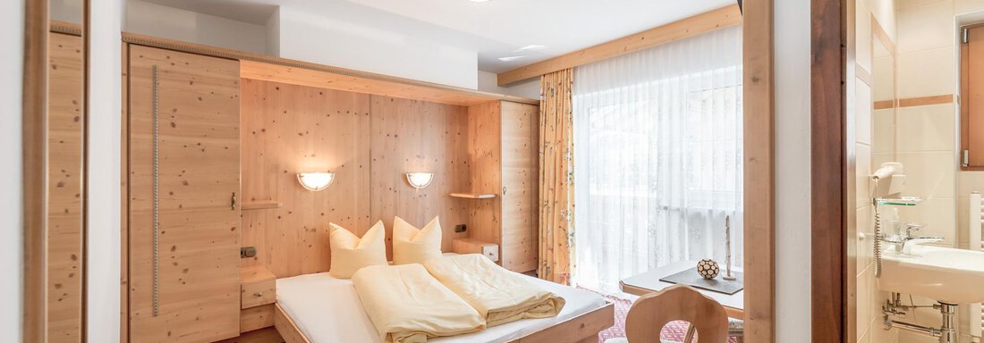 Schlafzimmer | © Sonnzeit (CC C) A cozy hotel room with a double bed and wooden interiors. There is a chair and a window with curtains that lets in daylight. - Ferienhaus Apart Sonnzeit - Sölden