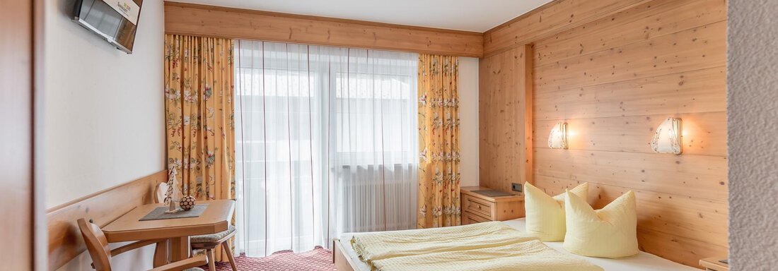 A cozy bedroom with wood paneling and a large window. The room is furnished with a double bed, a table, and a television. - Ferienhaus Apart Sonnzeit - Sölden