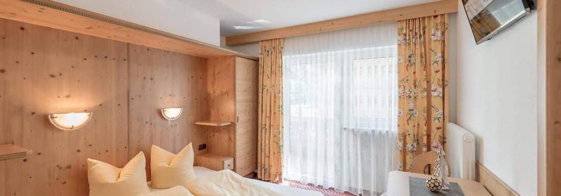 A cozy hotel room with wood paneling and a double bed. The room has large windows with curtains and a small desk. - Ferienhaus Apart Sonnzeit - Sölden