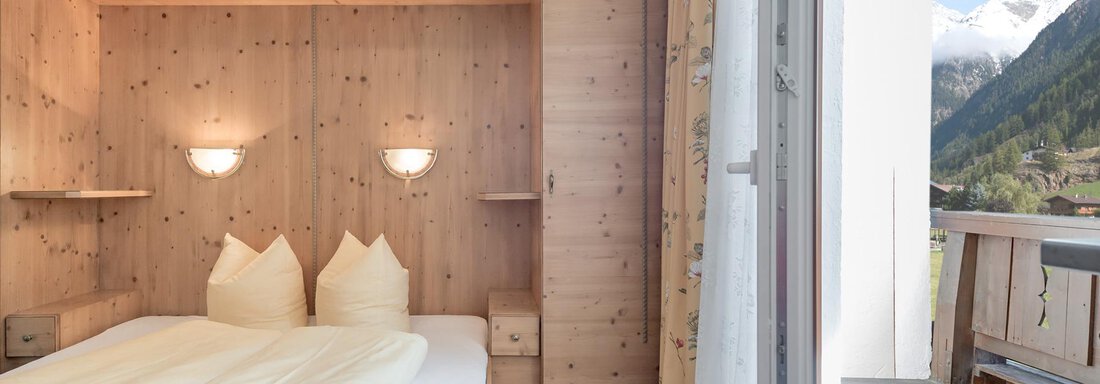 A cozy bedroom with wooden paneling and two pillows on the bed. The balcony offers a view of the surrounding mountains. - Ferienhaus Apart Sonnzeit - Sölden