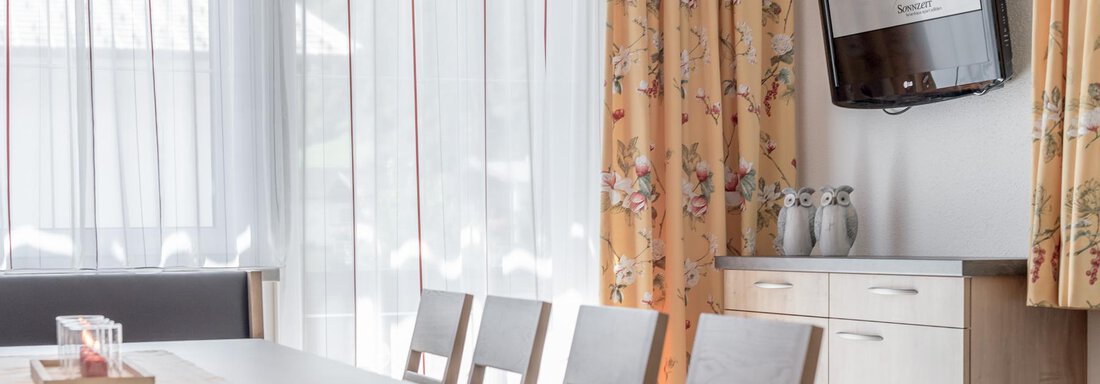 A bright dining area with a wooden table and chairs. There is a television on the wall, and the windows are adorned with light curtains. - Ferienhaus Apart Sonnzeit - Sölden