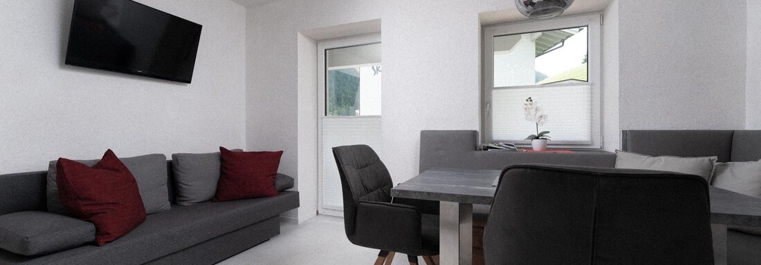 A modern living room with a gray sofa and a dining table. There is a television on the wall and bright windows that provide plenty of daylight. - Ferienhaus F2 - Sölden - Sölden