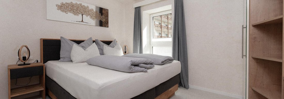A modern bedroom with a large bed and three pillows. The room has windows with curtains and a wooden wardrobe. - Ferienhaus F2 - Sölden - Sölden