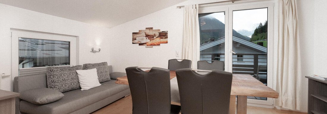 A modern living room with a gray sofa and a dining area. Large windows let in plenty of light and offer a view of the surroundings. - Ferienhaus F2 - Sölden - Sölden