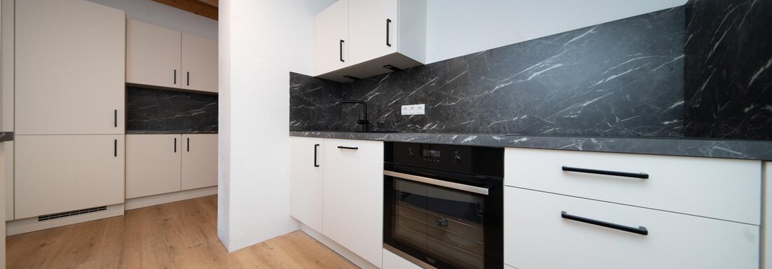 A modern kitchen with white cabinets and a black countertop. The floor is made of light wood and the walls are kept simple. - Ferienhaus F2 - Sölden - Sölden