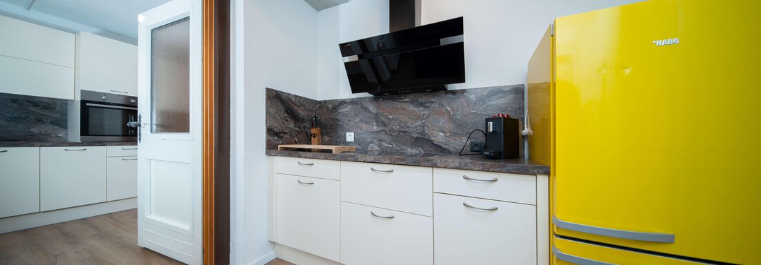 A modern kitchen with white cabinets and a yellow refrigerator. A black television hangs on the wall, and the work surface is made of gray material. - Ferienhaus F2 - Sölden - Sölden