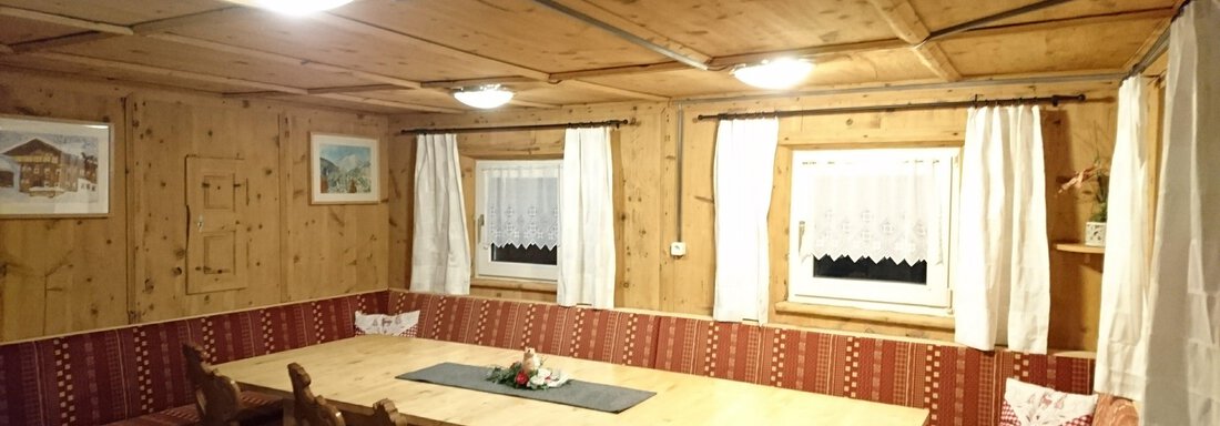 A cozy lounge area with wooden paneling and a large table. The windows are adorned with white curtains and the benches invite you to sit. - Ferienhaus Kaspers - Sölden