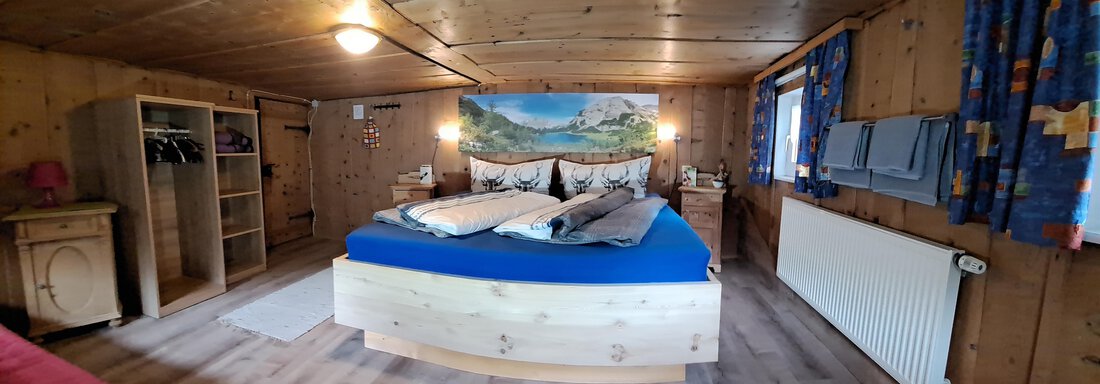 A cozy bedroom with a wooden ceiling and large windows. The centerpiece is a large bed with blue bedding and a picturesque mural. - Ferienhaus Kaspers - Sölden