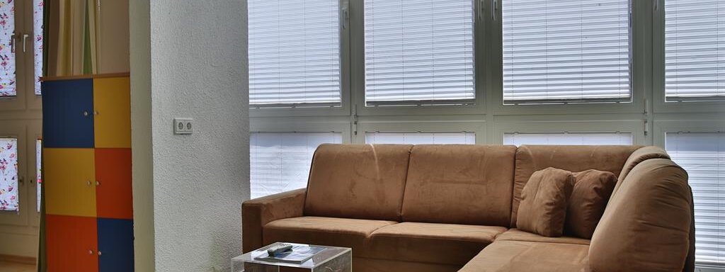 A cozy living room with a brown couch and a clear glass table. The windows are fitted with blinds and there is a colorful cabinet in the background. - Ferienhaus Moidl - Sölden