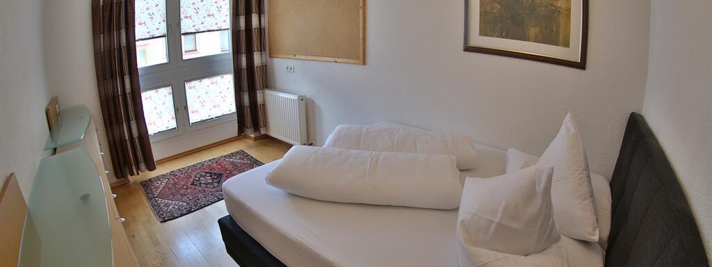 A simple bedroom with a bed, pillows, and a rug. The walls are light and there are large windows with curtains. - Ferienhaus Moidl - Sölden
