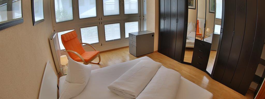 A bright bedroom with a large bed, an orange armchair, and a mirrored wardrobe. Large windows provide plenty of daylight. - Ferienhaus Moidl - Sölden