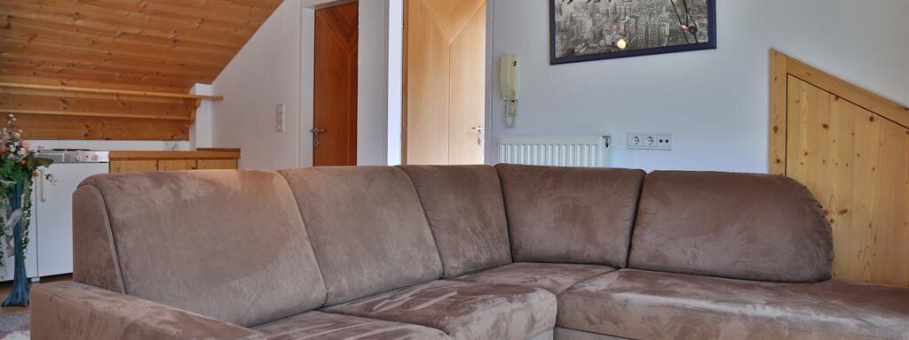 A cozy living room with a brown corner sofa and wooden ceilings. In the background, there is a picture on the wall and an open door visible. - Ferienhaus Moidl - Sölden
