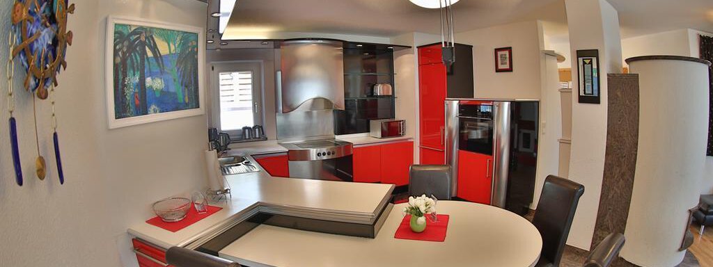 A modern kitchen with red cabinets and an oval dining table. The room design is bright and inviting with a view of the cooking area. - Ferienhaus Moidl - Sölden