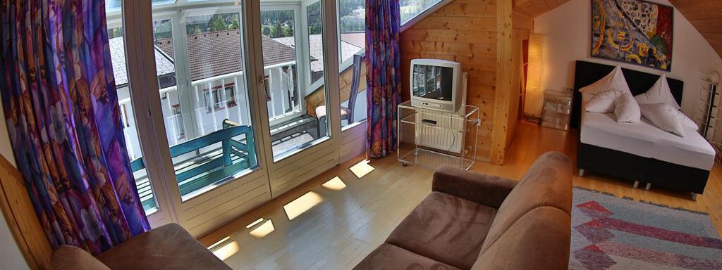 A cozy room with a modern sofa, large windows, and colorful curtains. The room features a television and a double bed, surrounded by wooden walls. - Ferienhaus Moidl - Sölden