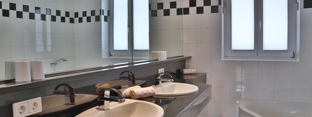 A modern bathroom with two sinks and a large mirror. The walls are tiled in white and black, and there is a bathtub in the corner. - Ferienhaus Moidl - Sölden