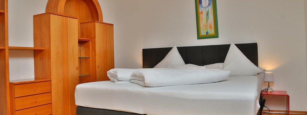 A bright, modern bedroom with a large bed and two pillows. A painted picture hangs on the wall, and there is a wooden wardrobe and a nightstand. - Ferienhaus Moidl - Sölden
