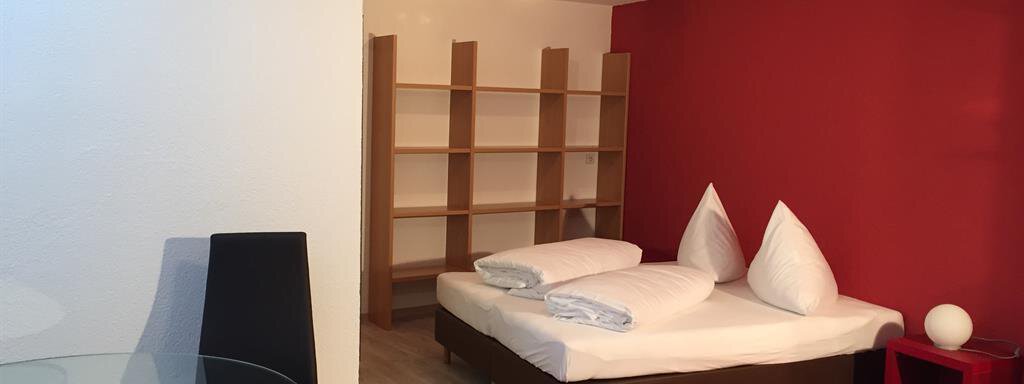 A modern room with a double bed and a red accent wall. There is a small table and chairs as well as shelves in the background. - Ferienhaus Moidl - Sölden