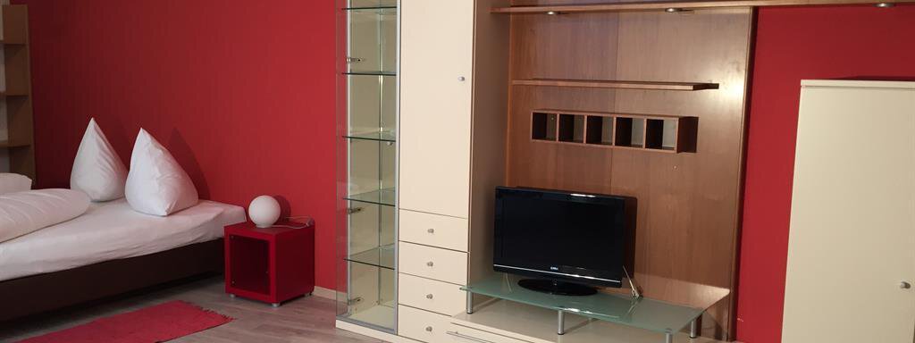 A modern living room with a red wall and a cozy bed. There is a TV unit and a small table next to a white light fixture. - Ferienhaus Moidl - Sölden