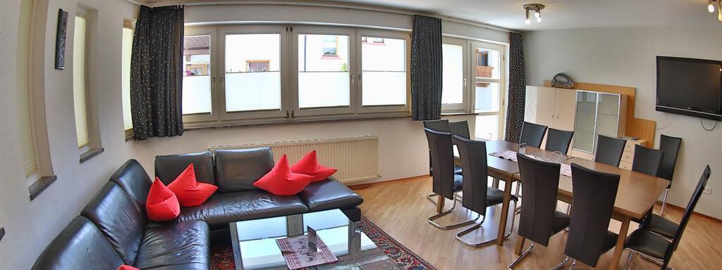 A modern living room with a cozy black couch and red cushions. There is a dining table with chairs and plenty of daylight coming through the windows. - Ferienhaus Moidl - Sölden