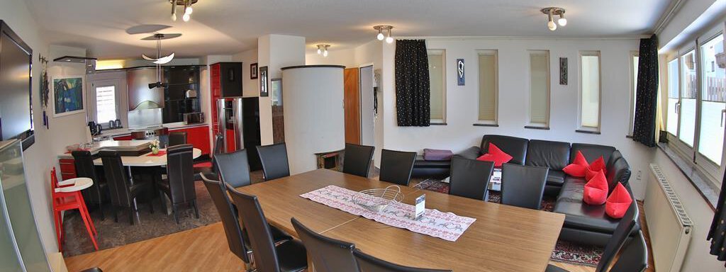 An open living area with a large dining table and modern black chairs. In the background, there is a cozy seating area and a kitchenette. - Ferienhaus Moidl - Sölden