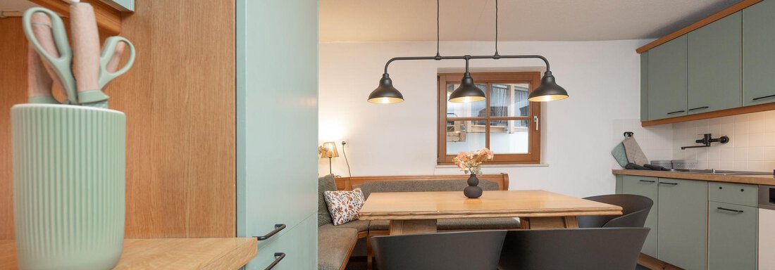 A modern kitchen with light green cabinets and a large dining table. There are cozy seating areas and attractive lighting. - Ferienhaus Valsora - Sölden