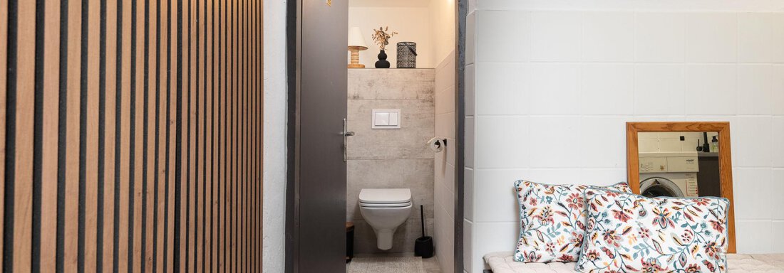A modern entrance area with a door leading to a bathroom. Decorative cushions are placed on a bench, and a mirror is attached to the wall. - Ferienhaus Valsora - Sölden