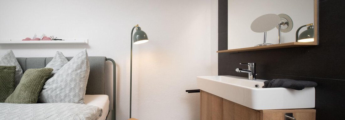 A modern bedroom with a large bed and cozy pillows. Next to the bed, there is a minimalist sink and a stylish floor lamp. - Ferienhaus Valsora - Sölden