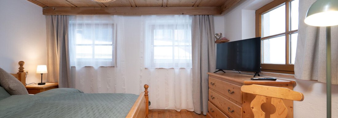 A modern bedroom with a double bed and wooden details. There is a dresser, a television, and large windows that let in plenty of light. - Ferienhaus Valsora - Sölden