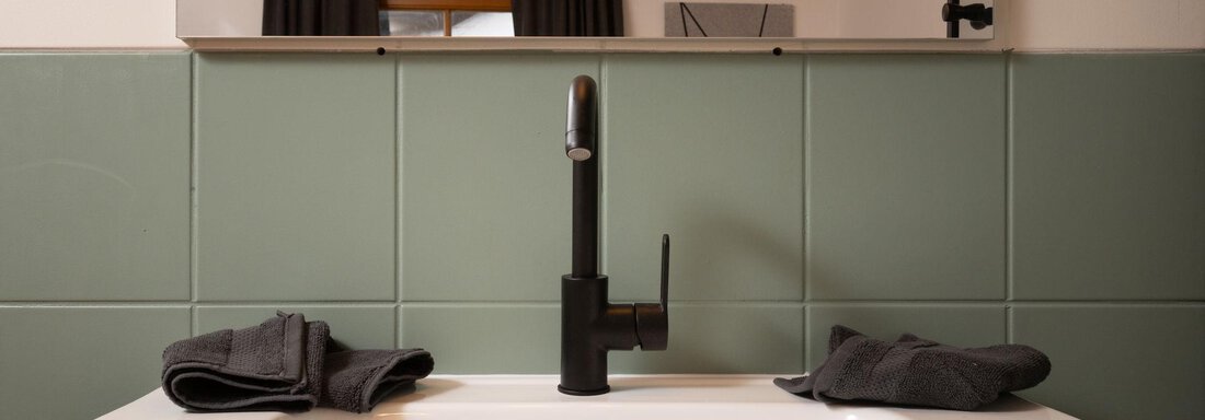 A modern sink with a black faucet and green tiles in the background. The bathroom atmosphere is expressed by a cozy lamp and towels. - Ferienhaus Valsora - Sölden