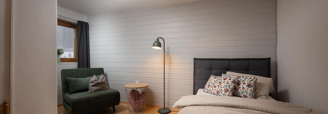 A cozy bedroom with a large bed and many pillows. There is an armchair, a side table, and warm light from a pendant lamp. - Ferienhaus Valsora - Sölden